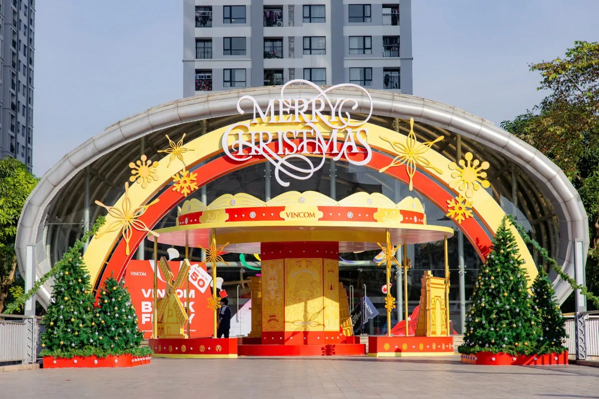 Cổng trang trí Merry Christmas lớn trước sảnh Vincom với cây thông Noel hai bên và các mô hình hộp quà màu đỏ vàng