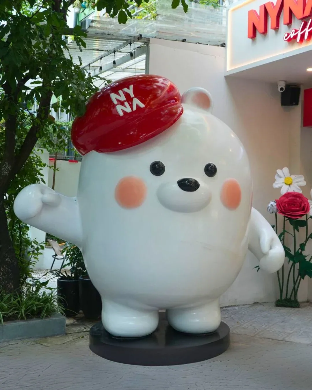 Mô hình mascot gấu trắng đội mũ đỏ NYNA bằng composite đặt trước cửa quán cà phê, cạnh hoa trang trí và bảng hiệu thương...