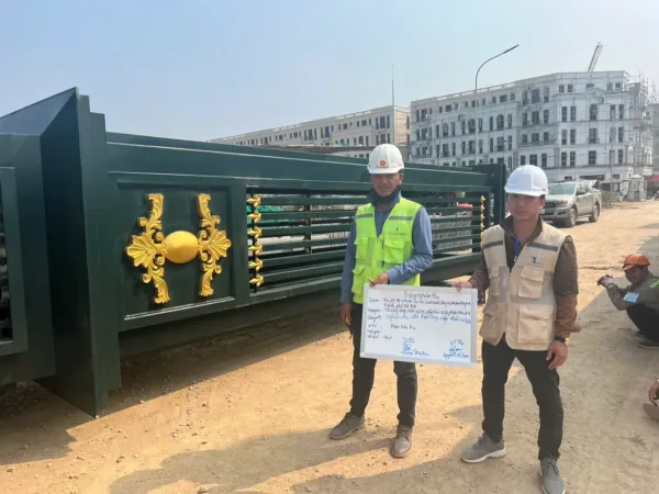 Hai nhân sự cầm bảng nghiệm thu trước cấu kiện cổng xanh đậm có phù điêu vàng.