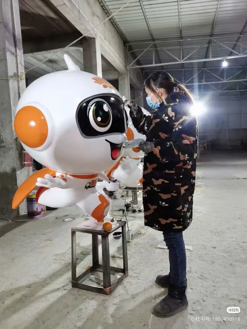 Thợ sơn đang hoàn thiện chi tiết trên mô hình linh vật robot màu cam trắng trong xưởng sản xuất.