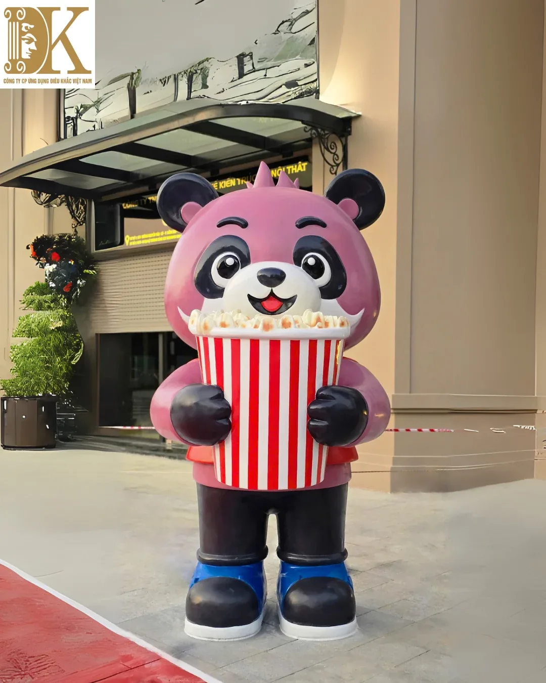 Mascot gấu trúc composite màu hồng ôm hộp bỏng ngô đỏ trắng đặt trước cửa showroom có mặt kính và bảng hiệu thương hiệu.